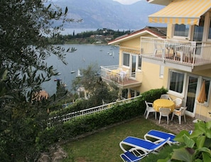 Ein zweistöckiges Gebäude mit einem Balkon mit Blick auf einen See mit Booten, ein kleiner Garten mit Tisch und Stühlen und ein blauer Liegestuhl.