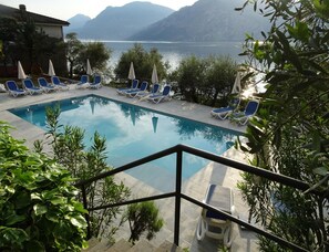 Ein Schwimmbecken mit Liegestühlen und Sonnenschirmen, umgeben von viel Grün und mit Blick auf ein Gewässer, im Hintergrund Berge.