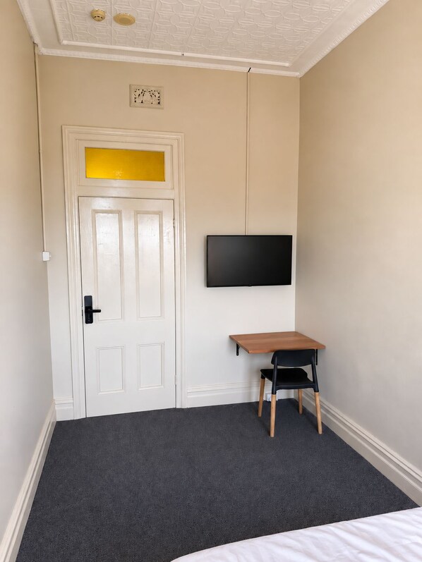 A room with a white door, a mounted flat-screen TV, a small wooden table, a black chair, and a dark carpeted floor.