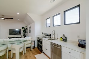 Una cocina moderna con comedor, que incluye una mesa de vidrio, sillas blancas, horno empotrado y pileta de acero inoxidable.