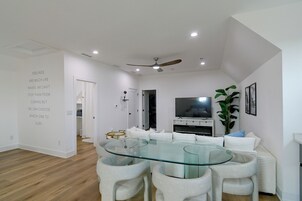 Una sala de estar moderna con una mesa de comedor de vidrio, sofás blancos, un ventilador de techo y un televisor de pantalla plana.