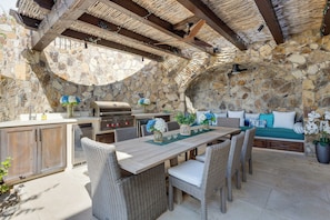 Cocina exterior con paredes de piedra, una mesa de comedor, sillas de mimbre y un área de descanso con cojines.