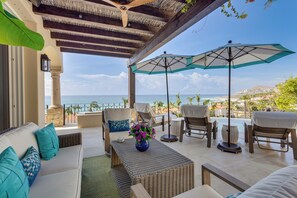 An outdoor seating area with a view of the sea, furnished with sofas and chairs, and a table with a vase of flowers.