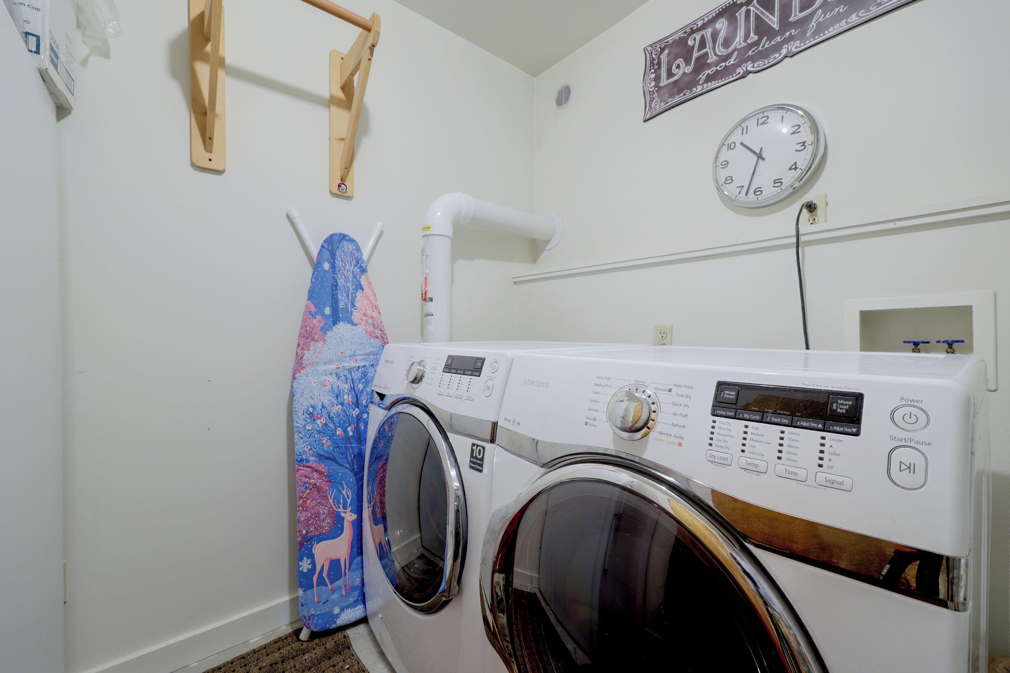 A laundry room with a front-loading washing machine, a wall-mounted clock, and a wooden drying rack.