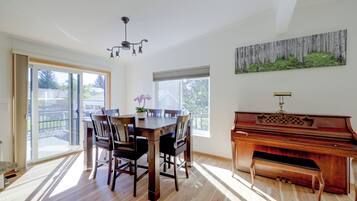 A dining area with a wooden table and chairs, a piano, and a large window with a view of the outdoors.