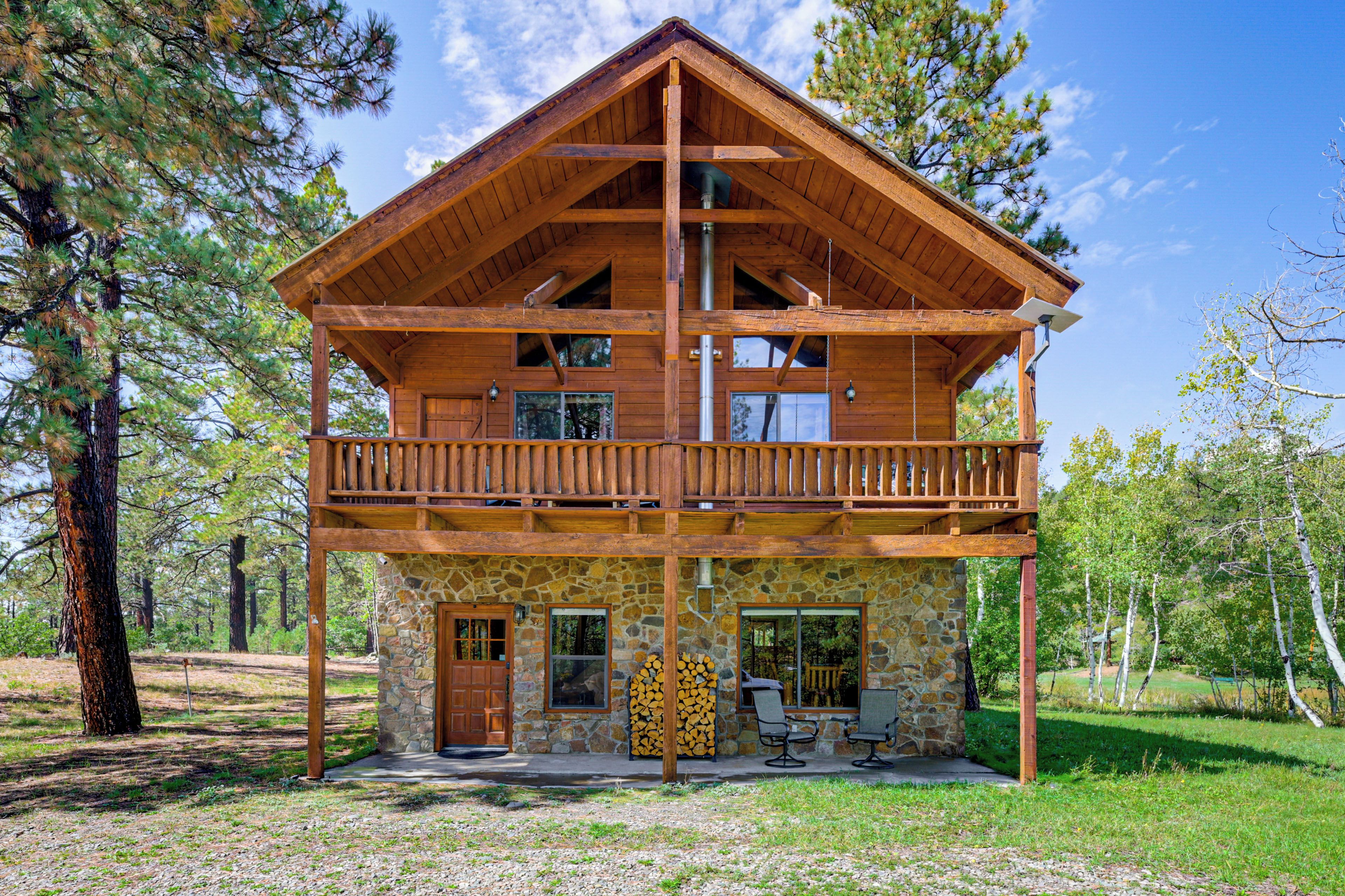 A two-story log cabin with a balcony, stone foundation, and outdoor seating area.