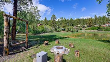 A natural landscape with a wooden swing set, a pond, and a stone fire pit.