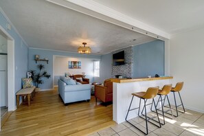 Un amplio salón con sofá, sillones y una mesa de comedor. Se ve una cocina con salpicadero de ladrillo y una barra.