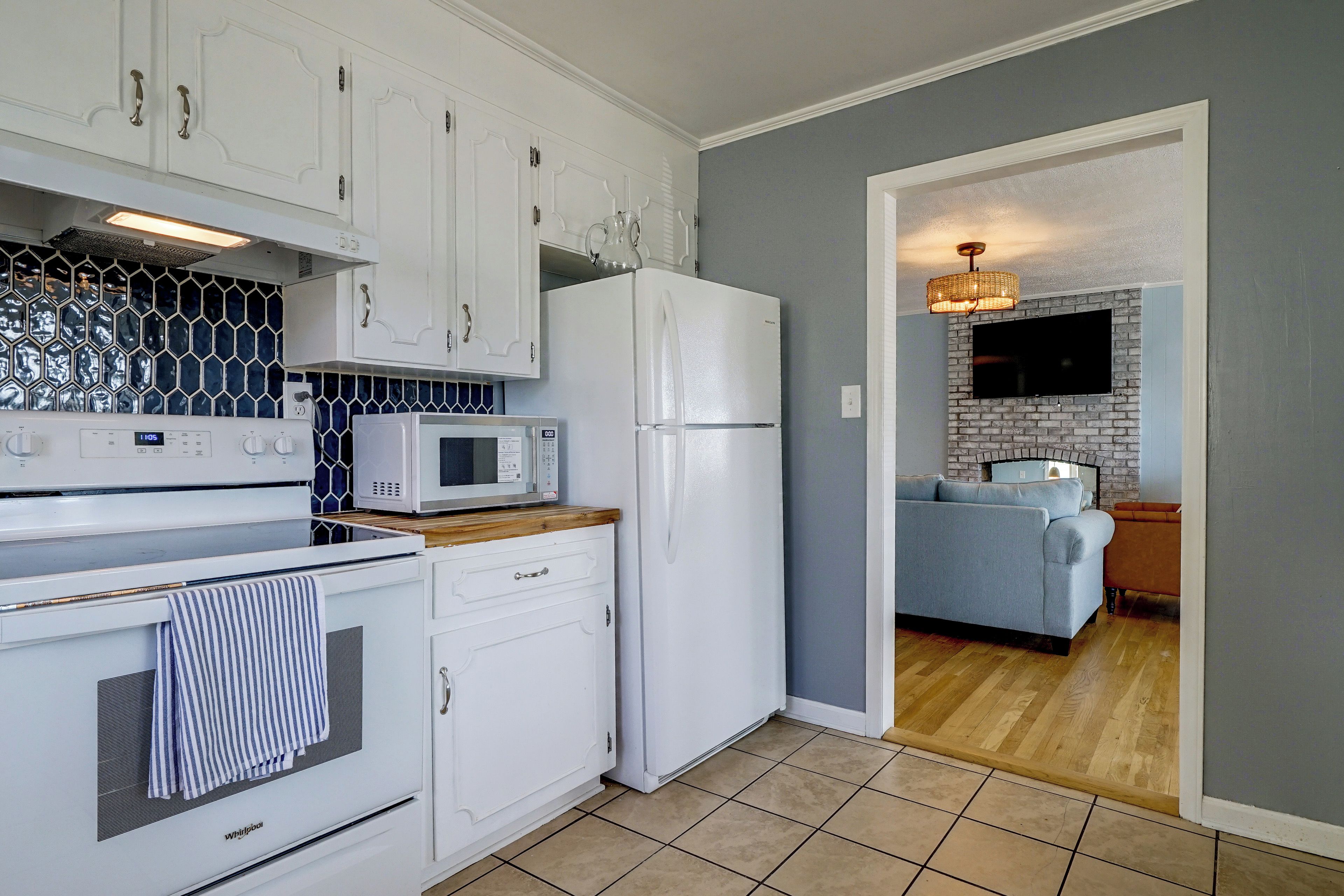 A compact kitchen with white appliances, a microwave, and a refrigerator. There is a wooden countertop and a striped towel hanging on the oven. The kitchen leads to a living area with a blue sofa and a television mounted on a brick wall.