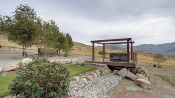 A rustic outdoor seating area with a wooden pergola, a grill, and a view of a hilly landscape.