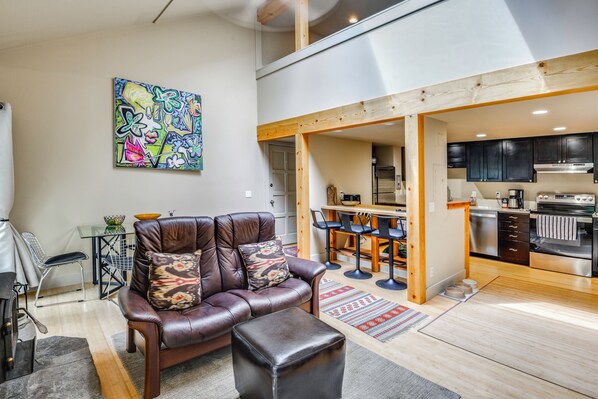 A modern living room with a leather sofa, a dining area with bar stools, and a kitchen with dark cabinets and stainless steel appliances.