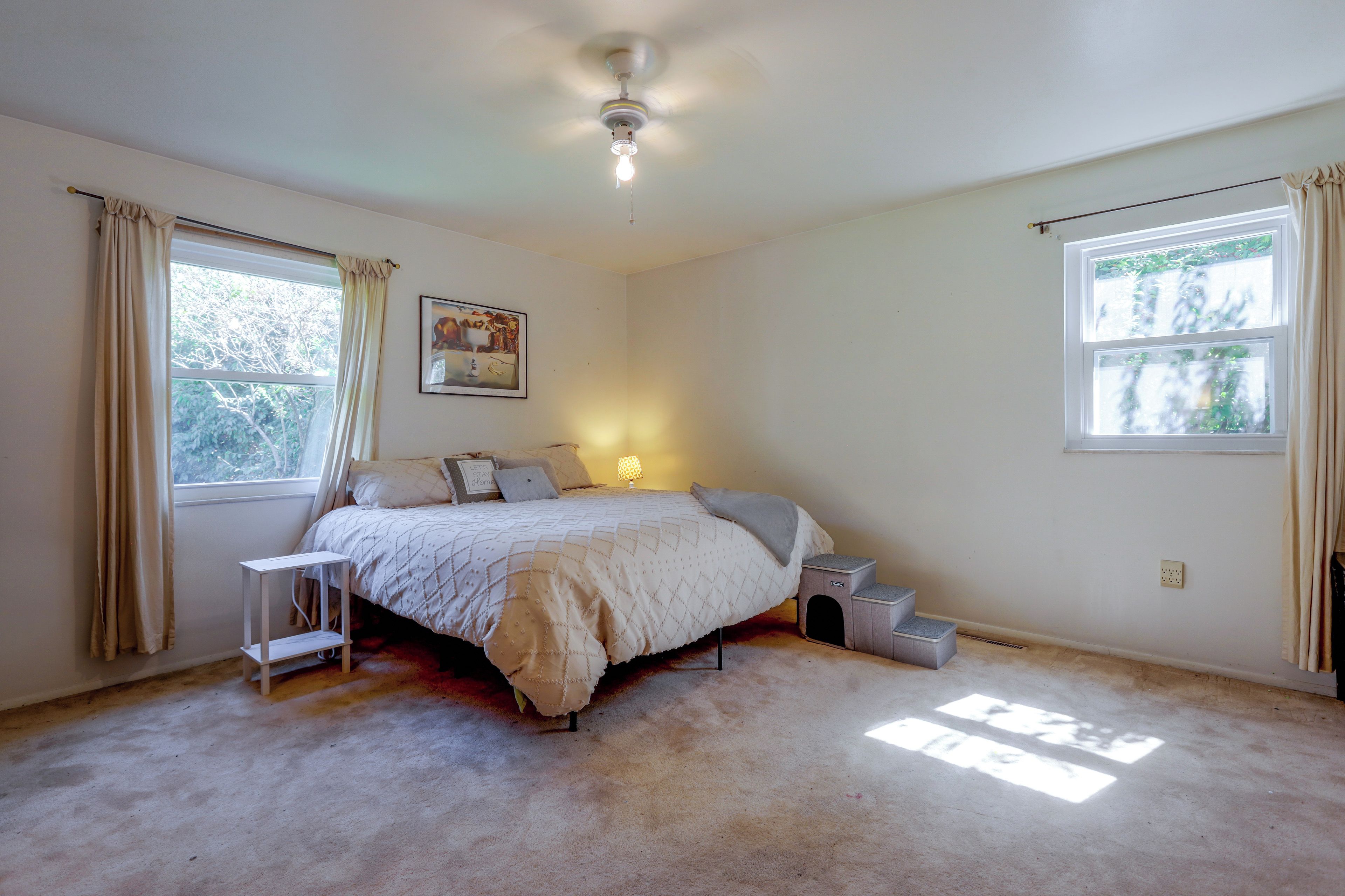 Une chambre à coucher avec un lit, une table de chevet, une lampe, une fenêtre et un ventilateur de plafond.
