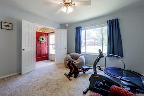 A room with a treadmill, a stationary bike, a red door, a white door, a ceiling fan, and a window with curtains.