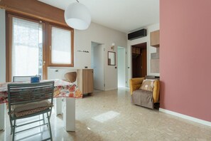 A spacious living area with a dining table, a sofa, and a chair. There is a window with curtains, a ceiling light, and a door leading to another room. - A cozy and bright 35 square-meter apartment, ideal for two people, located on the fifth floor of a building (elevator up to the third floor and is accessible only after a few steps; NOT suitable for people with disabilities) in a quiet, well-connected re (Milan)