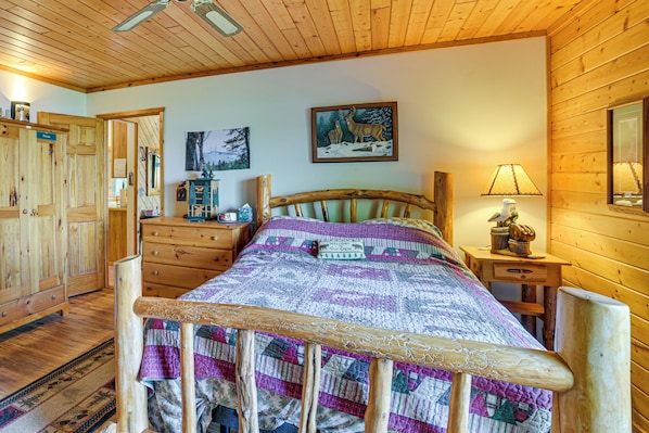 Interior de una cabaña con una cama de madera, un aparador, una mesita de noche y un cuadro en la pared.