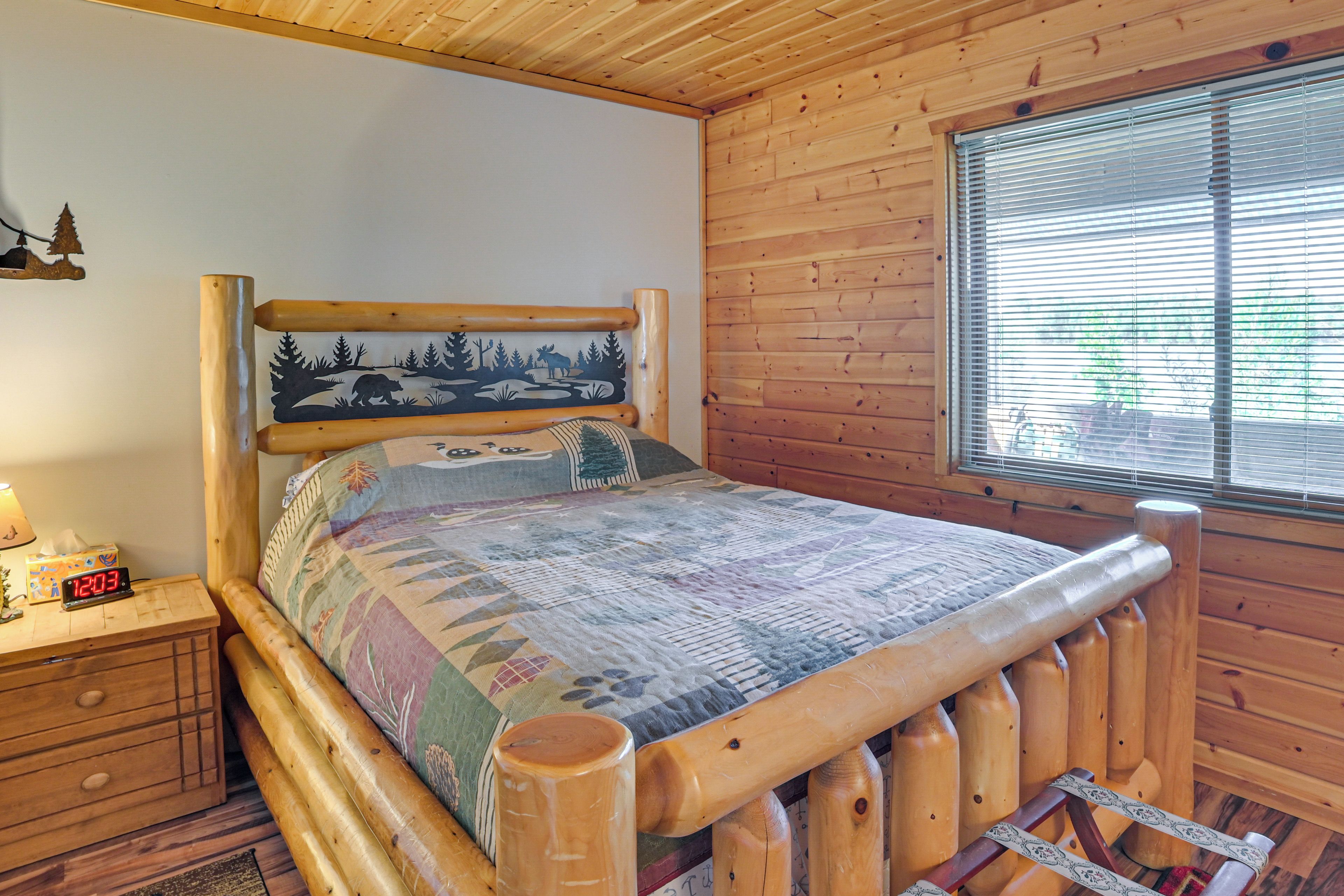 A wooden cabin room with a bed, a nightstand, a clock, and a window with blinds.