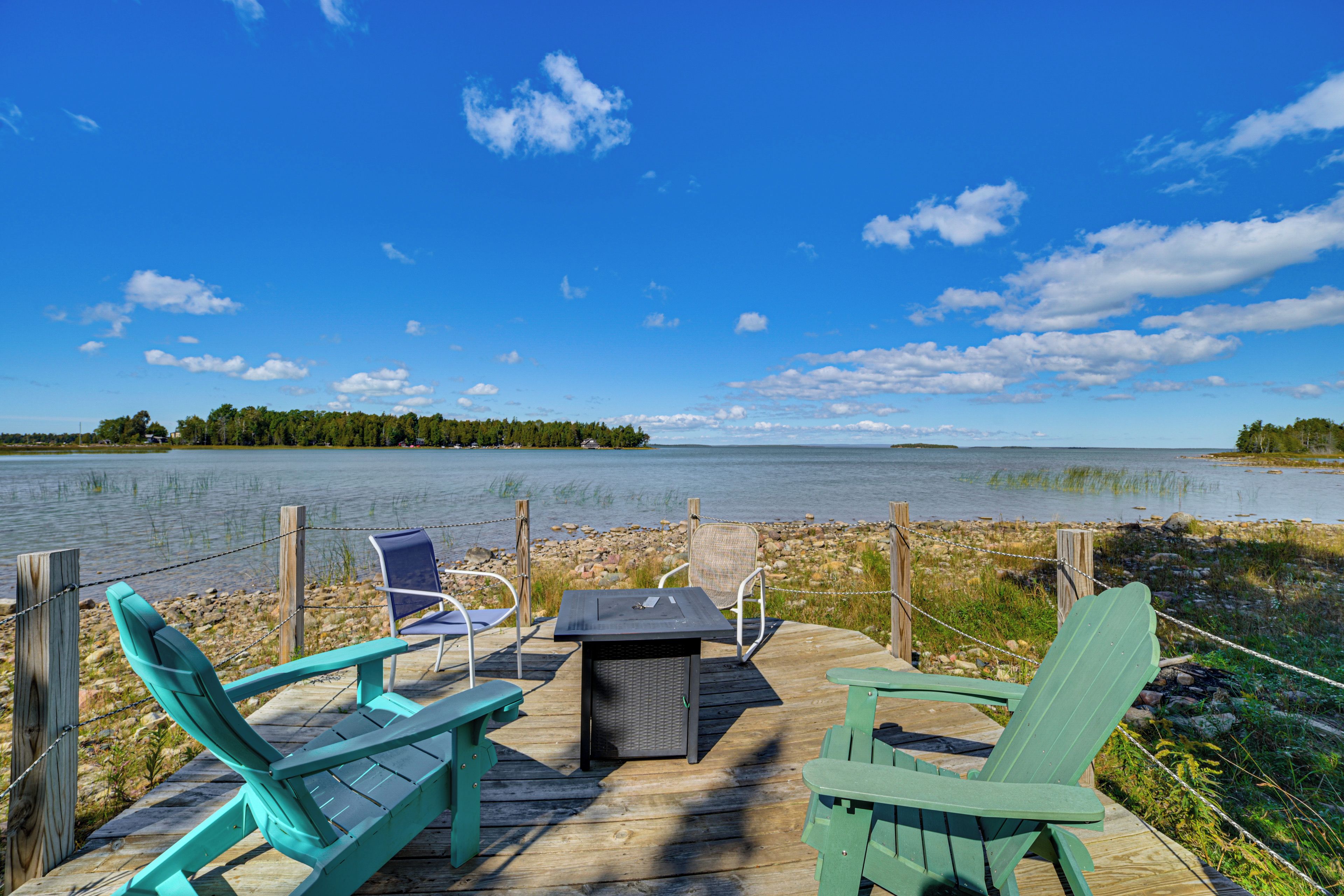 A wooden deck with two Adirondack chairs and a table, overlooking a calm lake with a forested shoreline.