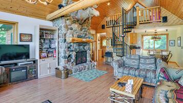 A spacious living room with a stone fireplace, a television, a bookshelf, and a cozy seating area with patterned cushions.