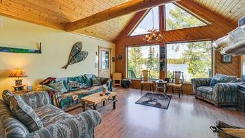 A cozy cabin interior with wooden floors, a large window offering a view of trees and a lake, and a fireplace.