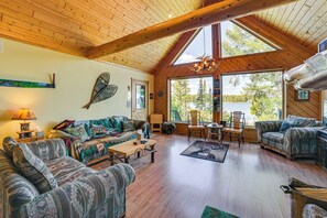 Interior acogedor de una cabaña con pisos de madera, un ventanal con vistas a árboles y un lago, y una chimenea.