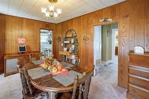 A dining room with a wooden table, chairs, a chandelier, and a kitchen in the background.