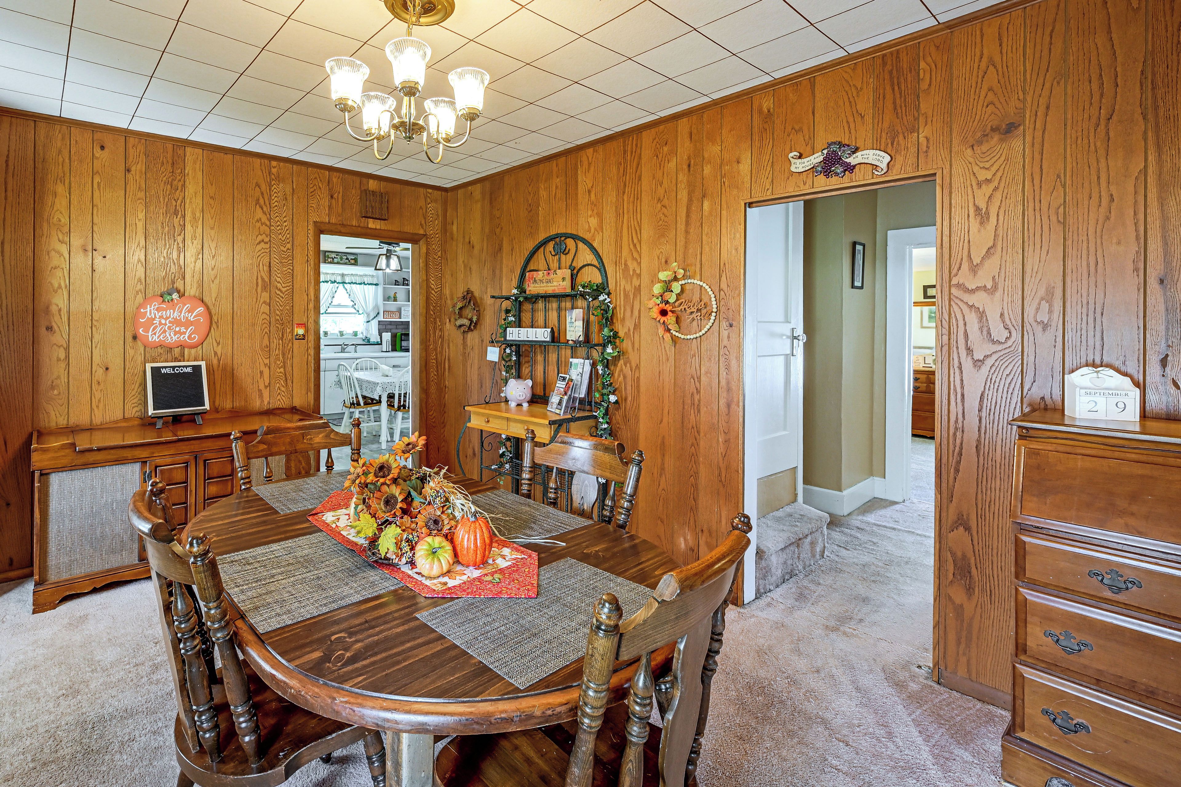 A dining room with a wooden table, chairs, a chandelier, and a kitchen in the background.