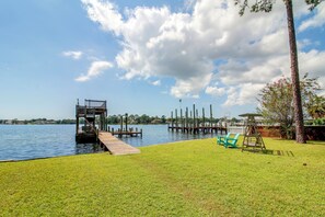 A grassy area with a dock extending into a lake, surrounded by trees and a clear sky. - Game Room & Hot Tub! Bayfront Family Gem (Fort Walton Beach)