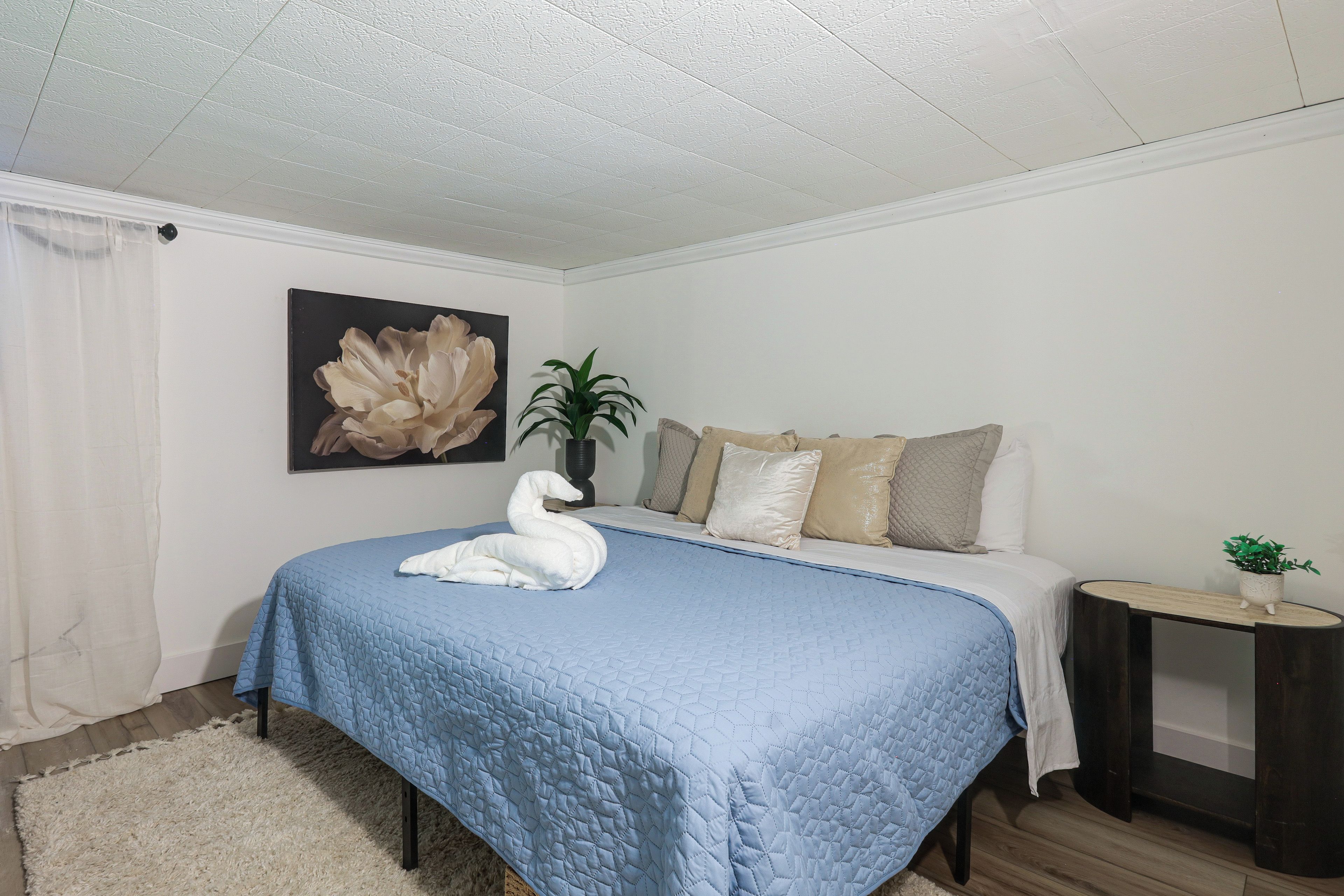 A bedroom with a bed, pillows, a bedside table, a plant, and a painting on the wall.