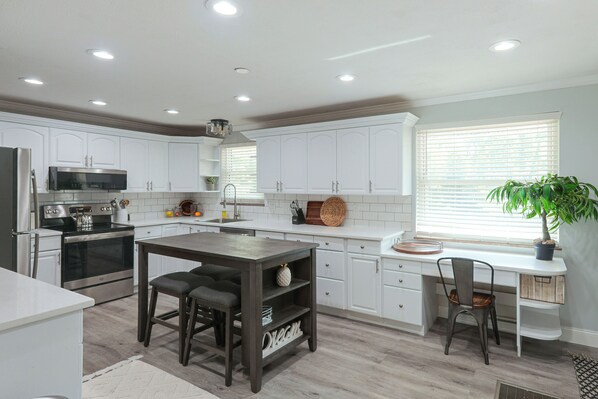 A modern kitchen with white cabinets, stainless steel appliances, a central island with stools, and a window with blinds.