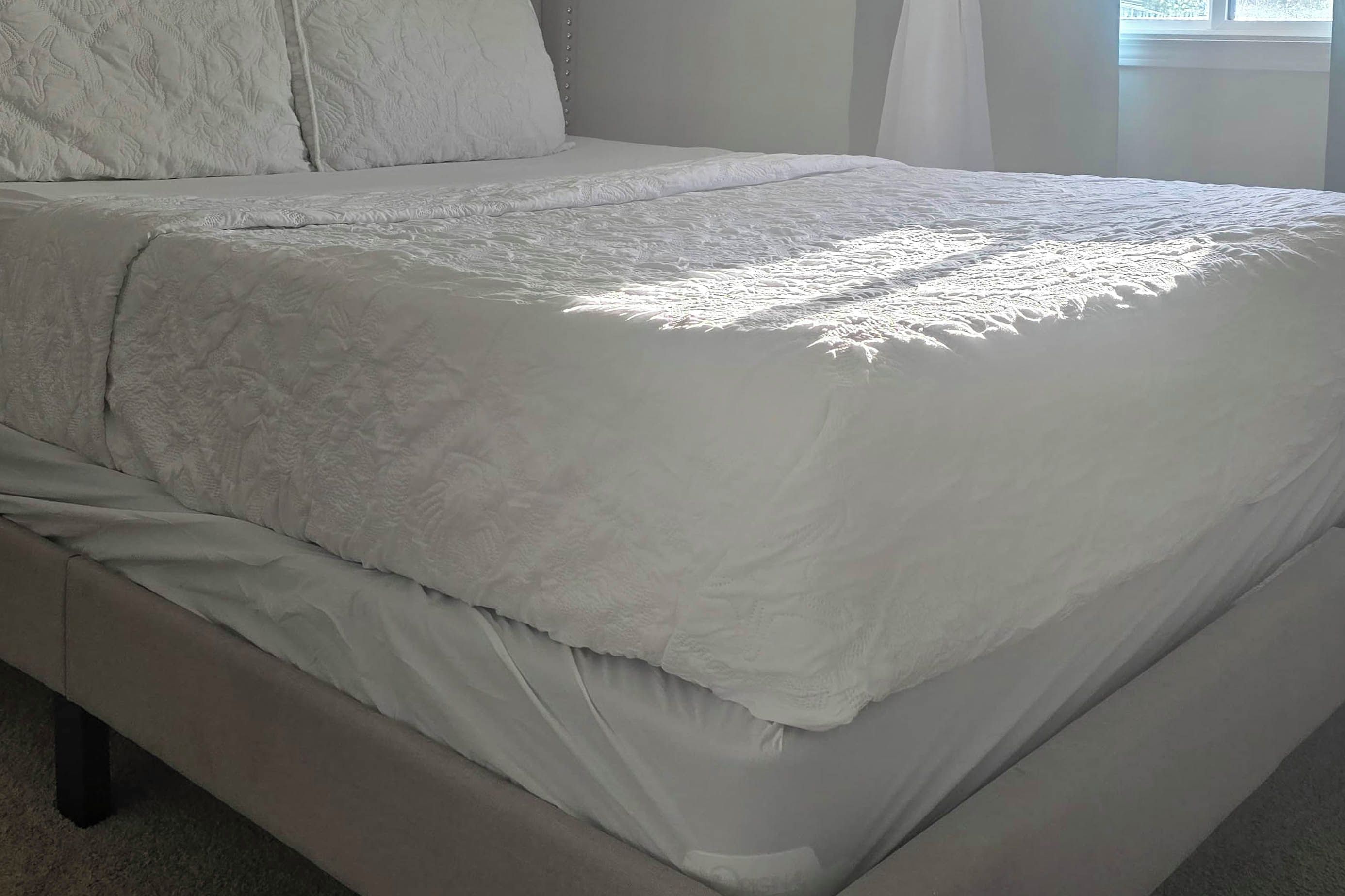 Una cama bien tendida, con una colcha blanca y almohadas, un cabecero y una ventana con cortinas.