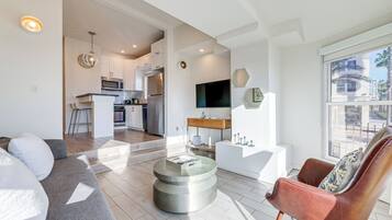 A modern living room with a sofa, a coffee table, and a leather armchair. There is a kitchen area with a microwave and a refrigerator in the background.