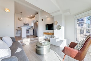 Una sala de estar moderna con un sofá, una mesa de centro y un sillón de cuero. Se observa un área de cocina con un microondas y un refrigerador al fondo.