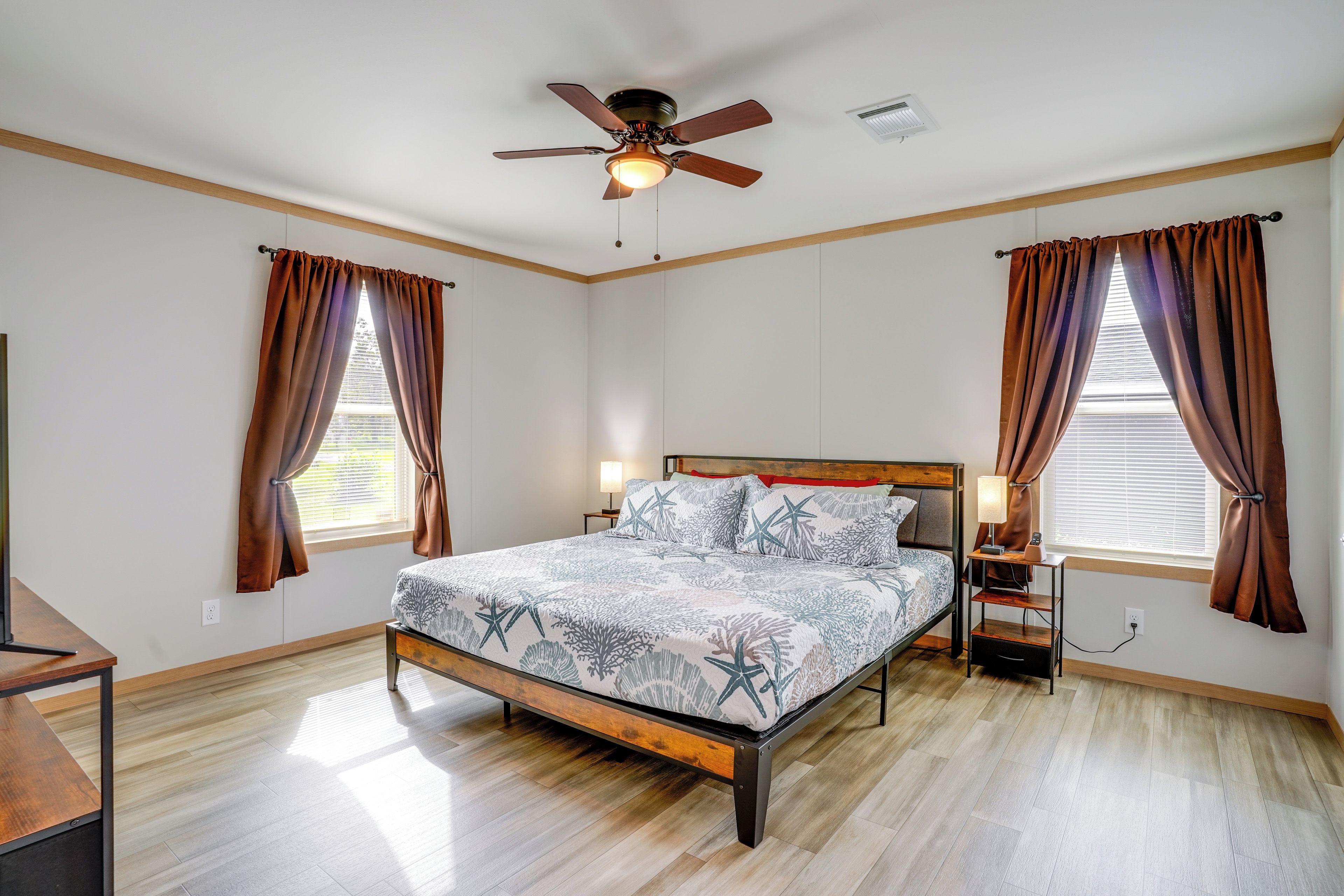 Un dormitorio con una cama, una mesita de noche, una lámpara, un ventilador de techo y dos ventanas con cortinas.