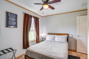 Un dormitorio con cama, ventilador de techo, ventana con cortinas, mesita de noche con lámpara y una frase motivacional en la pared.