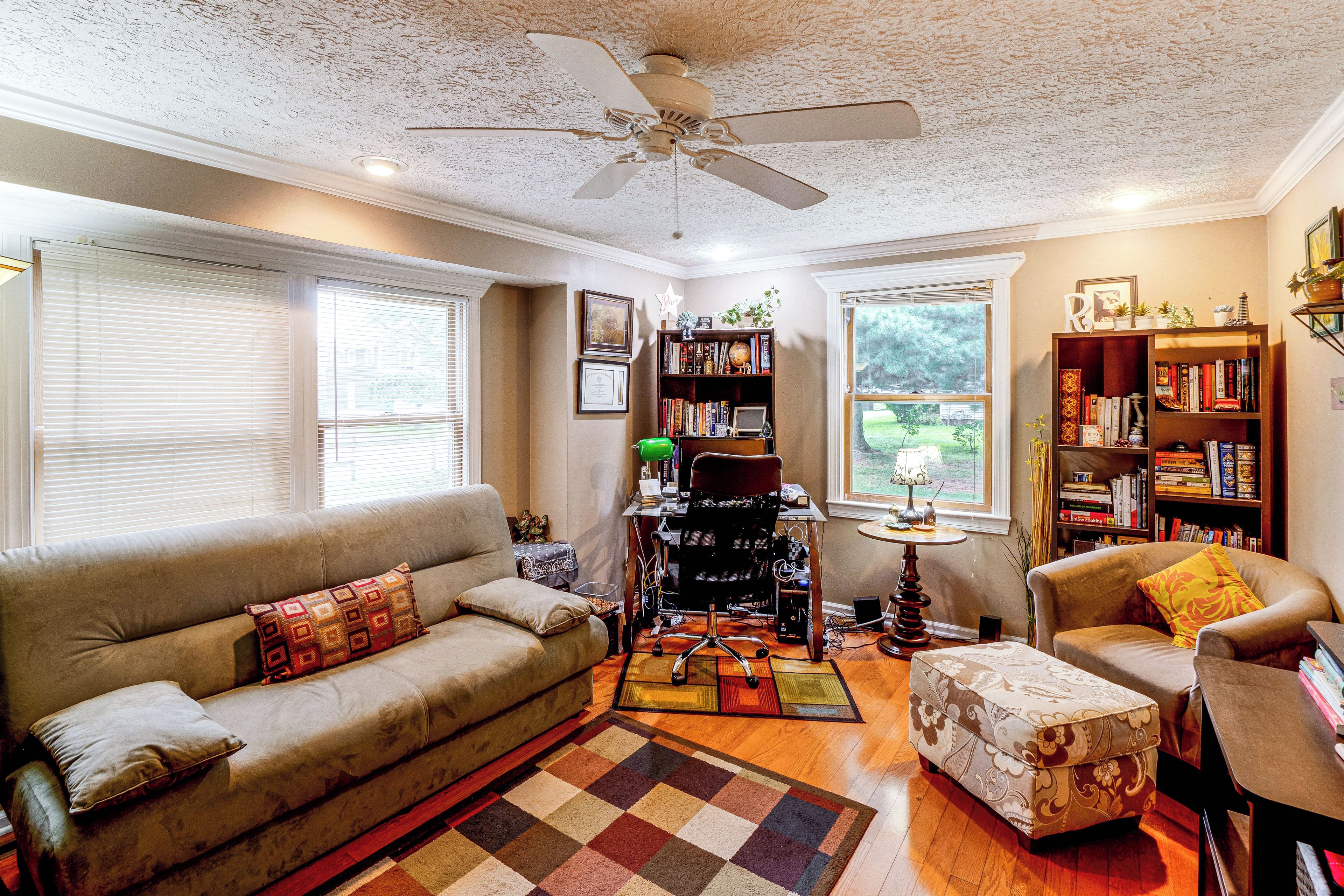 A living room with a sofa, armchair, and a desk with a computer. There are bookshelves and a ceiling fan.
