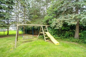 A wooden swing set with a slide in a grassy area surrounded by trees. - Explore Brockport! Family Gem, Neighborhood Perks (Brockport)