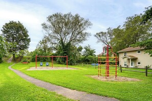 A playground with swings and a climbing structure, surrounded by grass and trees.
