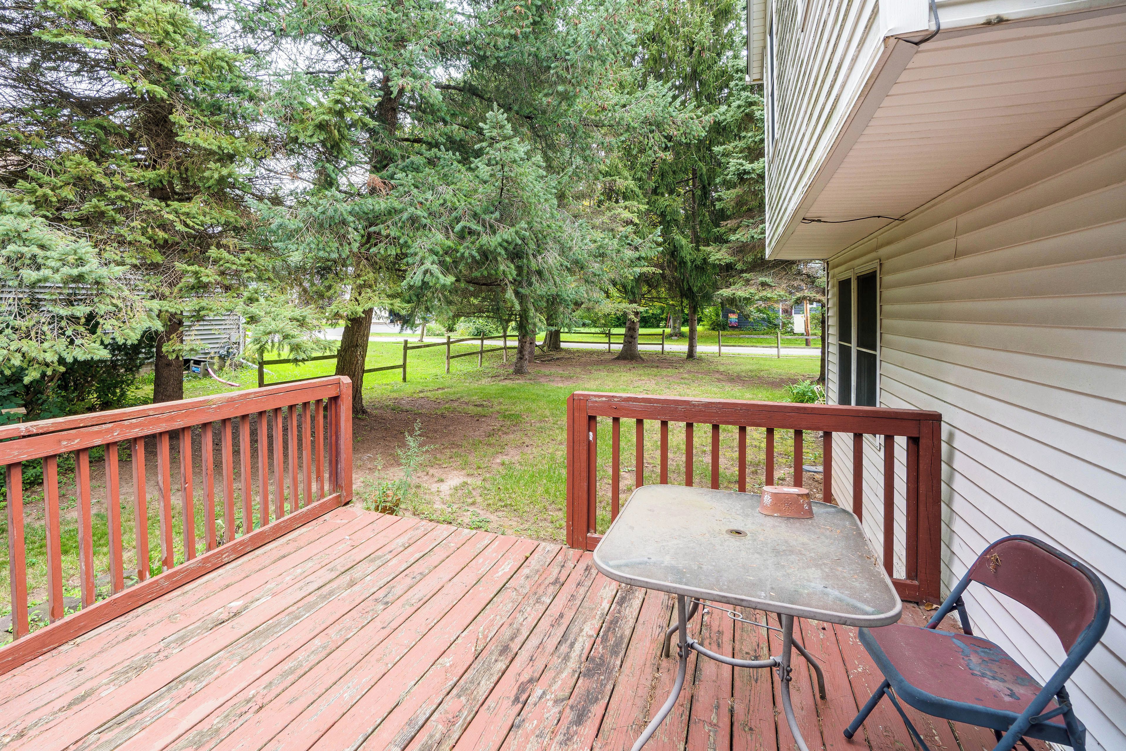 A wooden deck with a metal table and chairs, surrounded by trees and a grassy yard.