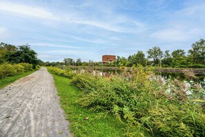Een verhard pad langs een waterpartij, met weelderige begroeiing aan één kant en een gebouw in de verte.