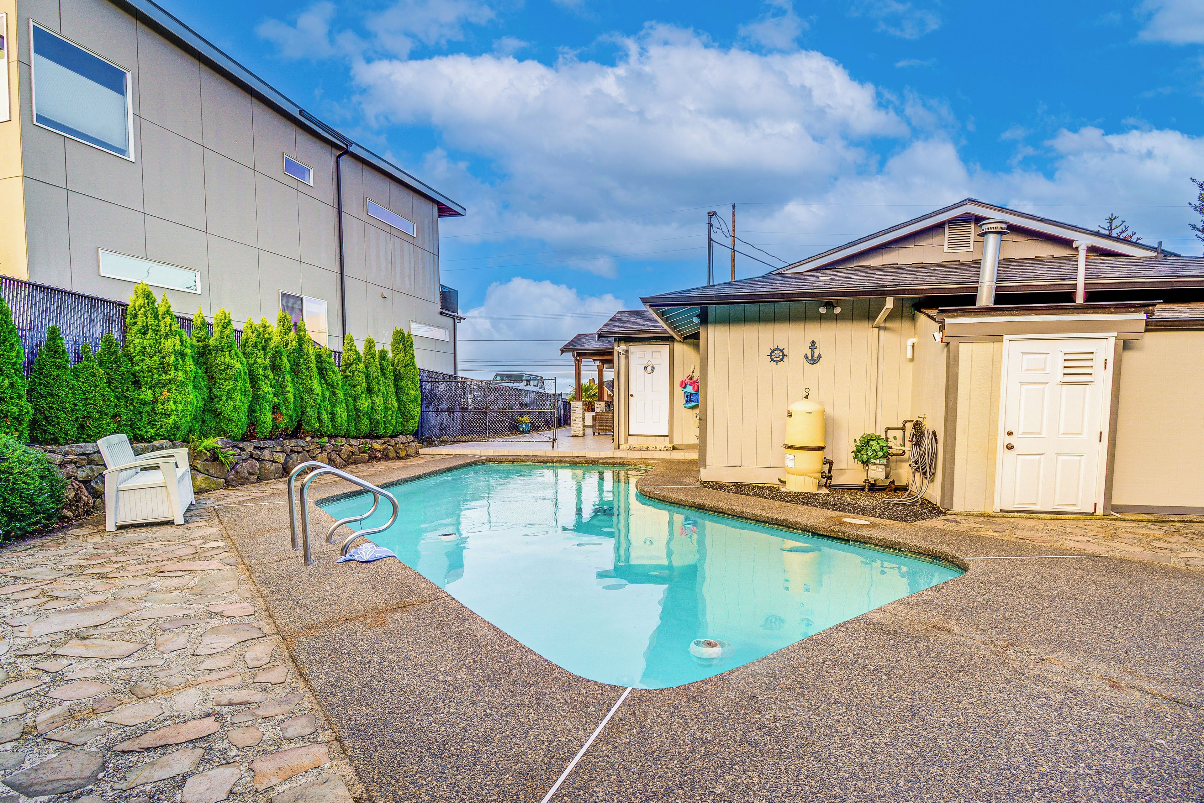 A swimming pool with a stone deck, a building with a grey facade, and a fenced area with greenery.
