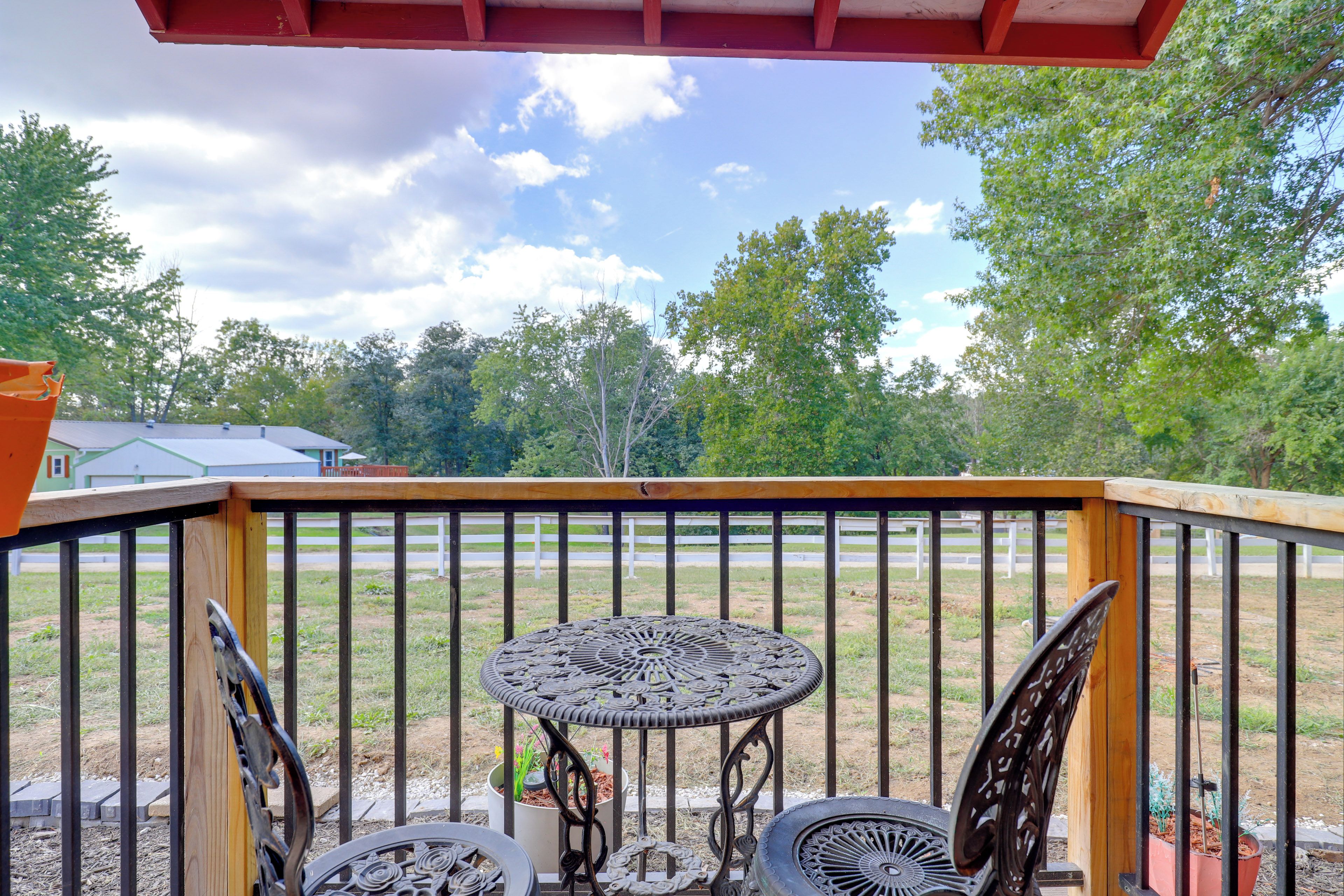 A wooden deck with a metal railing, a round table with chairs, and a view of a grassy field and trees.