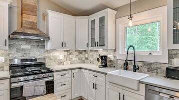 A modern kitchen with white cabinetry, a built-in stove, a coffee maker, and a window with a view of trees.