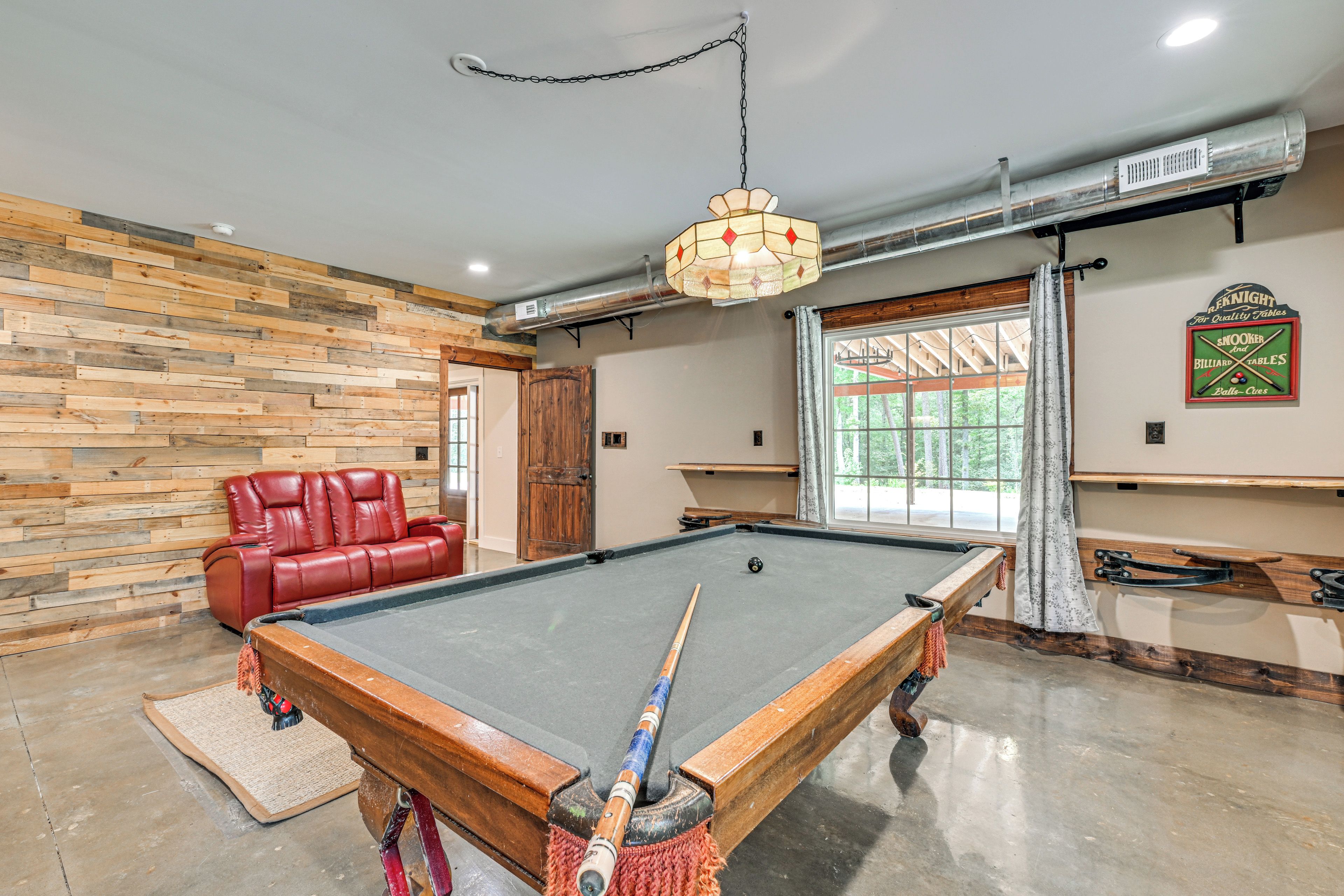 A pool table in a room with a wooden wall, a red leather sofa, and a chandelier.