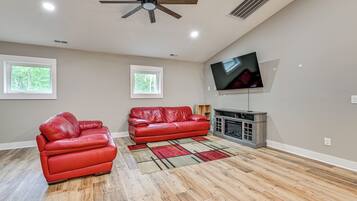 A spacious living room with a fireplace, two red leather sofas, a large flat-screen TV mounted on the wall, and a wooden ceiling fan.