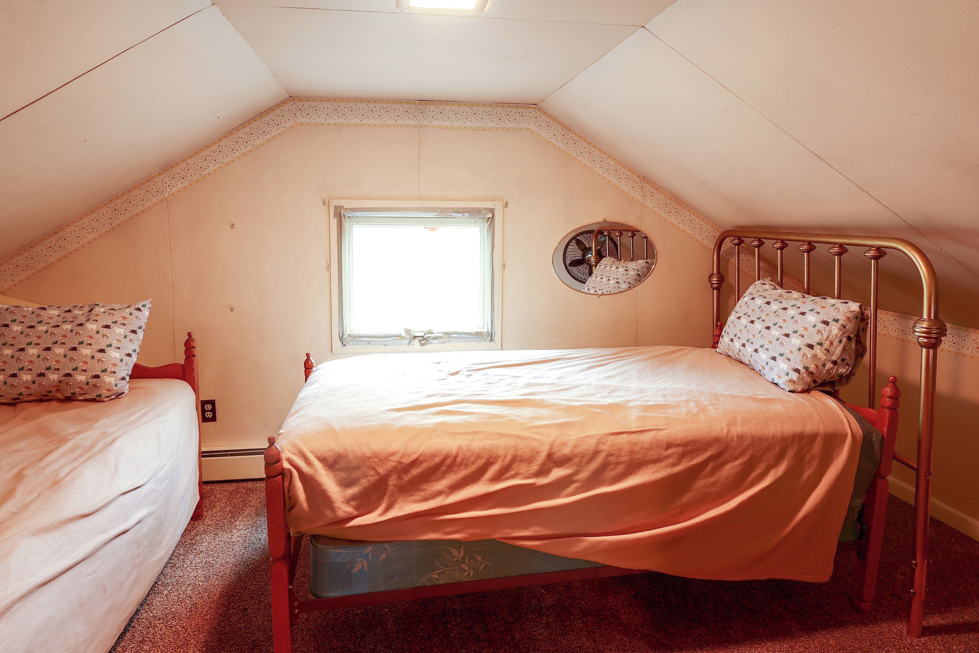 A small, single-bed room with a sloped ceiling, a window, and a wall-mounted fan.
