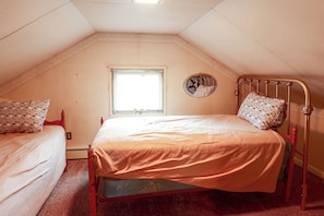 A small, single-bed room with a sloped ceiling, a window, and a wall-mounted fan.