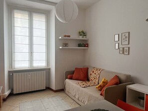 Ein Wohnzimmer mit einer Couch, einem kleinen Tisch, einer Heizung, einem Fenster mit Jalousien, einer Hängelampe und Wandregalen mit Dekorationen.