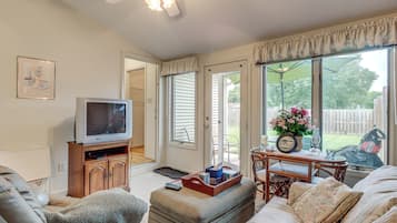 A living room with a television, sofa, ottoman, and a table with a clock and flowers.