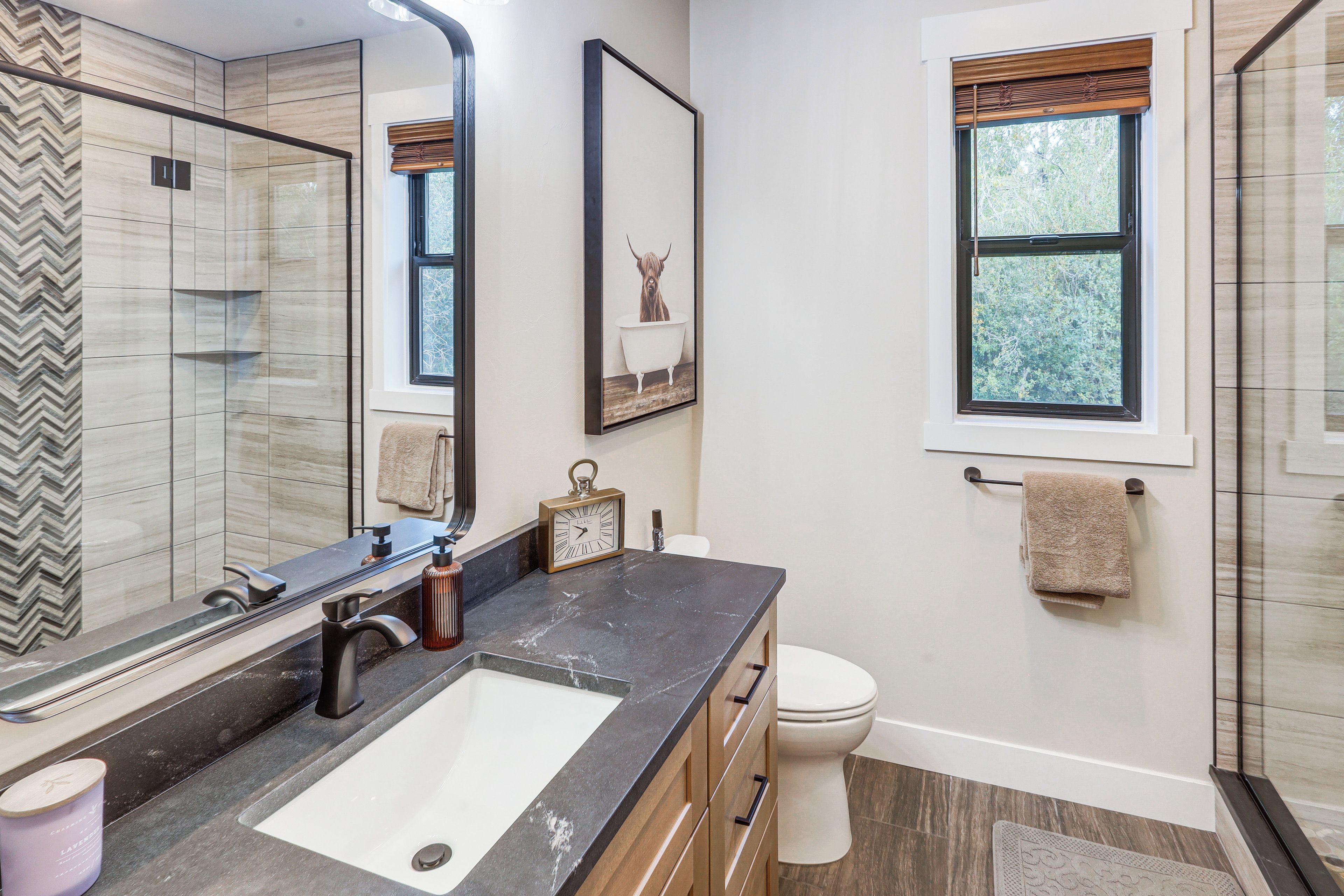A modern bathroom with a large mirror, a sink with a faucet, a toilet, and a window with a view of trees.