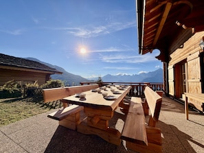 Ein Holztisch im Freien mit Bänken, vor einer Bergkulisse und blauem Himmel.